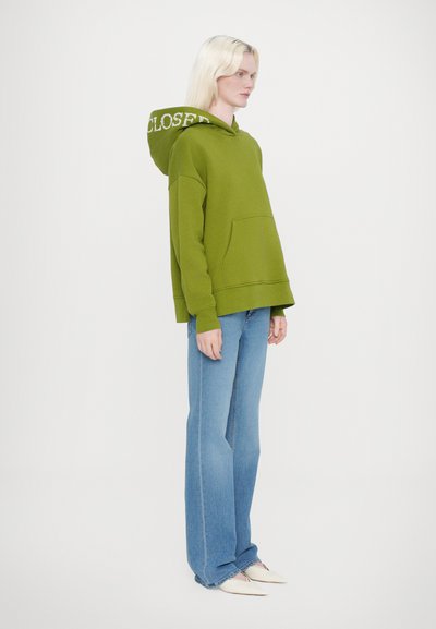 Grön huvtröja med ficka fram och texten "STÄNGD" på huvan, i kombination med ljusblå vida jeans och bleka skor.