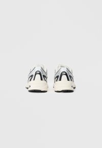 Une paire de chaussures de course blanches et noires vue de l'arrière sur un fond gris clair uni.