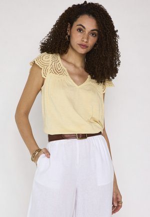 Donna con capelli ricci che indossa un top giallo con pizzo, senza maniche, pantaloni bianchi, cintura marrone e gioielli dorati, in piedi con una mano in tasca.