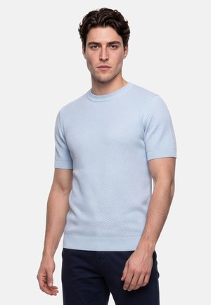 Junger Mann mit kurzen lockigen braunen Haaren, trägt ein hellblaues, strukturiertes Kurzarmhemd und dunkle Hose, steht vor weißem Hintergrund.