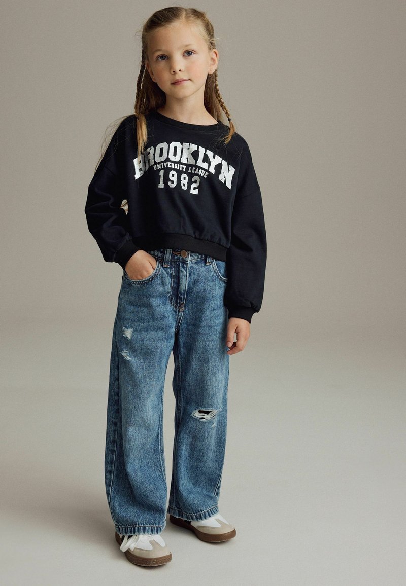 Sweatshirt noir court avec le texte "Brooklyn 1982", associé à un jean décontracté bleu clair usé et des baskets blanc-marron.
