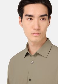 Korte mouwen, olijfgroene overhemd met knopen en een puntige kraag. Heeft een gladde textuur en een placket met twee knopen.