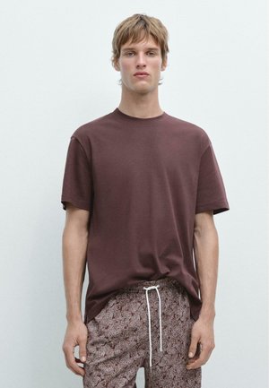 Jeune homme portant un t-shirt marron uni à manches courtes et un pantalon à cordon de serrage à motifs, debout devant un fond gris clair.