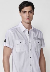 Camisa blanca de manga corta con cuello button-down, que presenta dos bolsillos en el pecho, botones negros y detalles de texto impreso en el lado izquierdo.