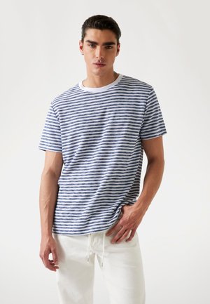 T-shirt à manches courtes rayé en blanc et bleu marine, en matériau coton avec un col rond et une coupe décontractée. Le modèle se tient avec les mains dans les poches.