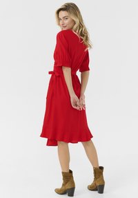 Robe rouge, longueur genou, avec des manches bouffantes courtes, une taille ceinturée et un ourlet à volants. Assortie à des bottines beige. Tissu lisse.