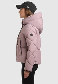 Prešívaná puffová bunda svetloružovej farby s kapucňou, prednými zipsovými vreckami a čiernym logom na rukáve. Skombinovaná s čiernym klobúkom.