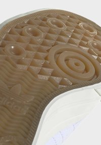 Suela de zapatilla de goma en beige con un patrón triangular texturizado. Murillo lateral blanco con sutiles estrías. Detalle circular en relieve notable.