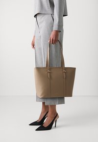 Sac fourre-tout en cuir synthétique beige avec deux poignées, présentant une surface texturée et des accents de ferrures dorées ; assorti à une tenue à carreaux et des talons noirs.