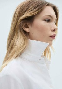 White collared shirt with a structured, high collar. The fabric appears smooth and lightweight, featuring subtle stitching and a button closure.