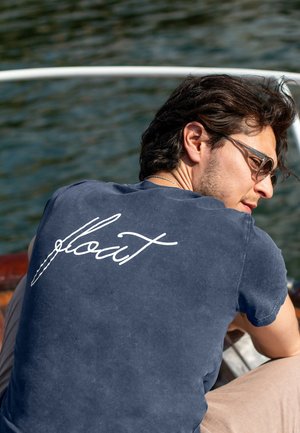 T-shirt in cotone blu navy con una texture morbida. Presenta il testo in corsivo bianco "float" sul retro. Maniche corte e vestibilità classica.