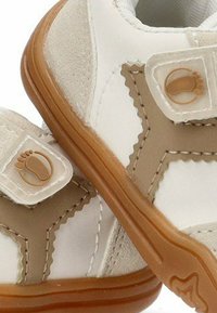 Zapatos de niño pequeños en color beige y blanco, con suelas de goma marrón y cintas de velcro que presentan un pequeño icono de pie.