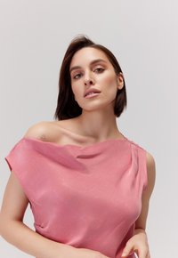 Różowa jedwabna bluzka o asymetrycznym dekolcie i lekko drapowanym fasonie. Gładka faktura i krótkie rękawy; posiada subtelny detal na ramieniu.