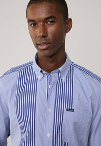 Camisa de botones a rayas azul y blanca con un bolsillo, que presenta un diseño de panel contrastante y una pequeña etiqueta de la marca en el pecho.