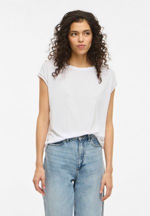 Kvinde med krøllet mørkt hår iført en enkel hvid kortærmet top og lyseblå højtaljede jeans, stående foran en hvid baggrund.