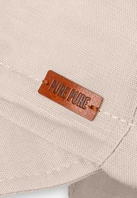 Beige caps laget av stoff med en teksturert overflate; har et lite brunt lærmerke merket "PURE PURE." Enkel, ren design.