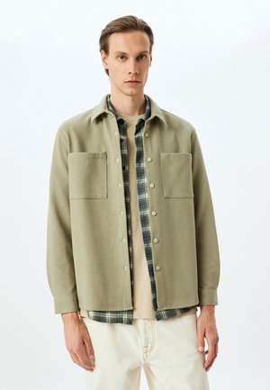 Chaqueta verde claro, de botones, con dos bolsillos en el pecho sobre una camisa de franela a cuadros, combinada con una camiseta beige y pantalones crema.