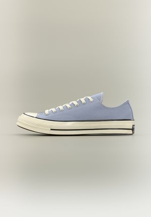 Zapatilla de lona azul claro de corte bajo con cordones blancos, suela de goma color crema y detalles de rayas negras, mostrada de perfil sobre un fondo neutro.