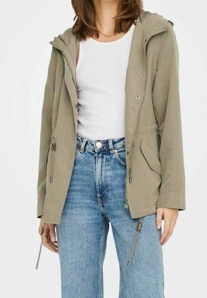 Person trägt eine beige Kapuzenjacke über einem weißen Trägertop und hoch taillierte blaue Jeans, steht vor einem schlichten Hintergrund.