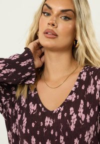 Vestido de tela acanalada con estampado floral en marrón oscuro con flores rosas. Presenta un escote en V profundo y mangas largas con volantes. Collar de cadena dorada.