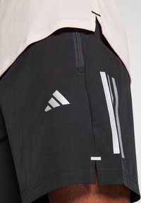 Μαύρες αθλητικές βερμούδες με ασημί λεπτομέρειες και λογότυπο Adidas, διαθέτοντας τσέπη με φερμουάρ και πλαϊνές σχισμές για καλύτερη κινητικότητα.