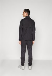 Man die met zijn rug naar ons toe staat, gekleed in een zwarte lange mouwen jas en zwarte broek, met witte sneakers, tegen een effen lichte achtergrond.