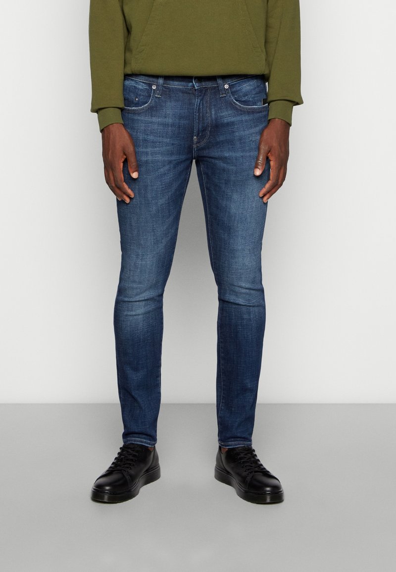 Slim-fit blå jeans i denimtyg med midjehöjd, femficksdesign, lätt bleknad yta, kombinerade med svarta sneakers.