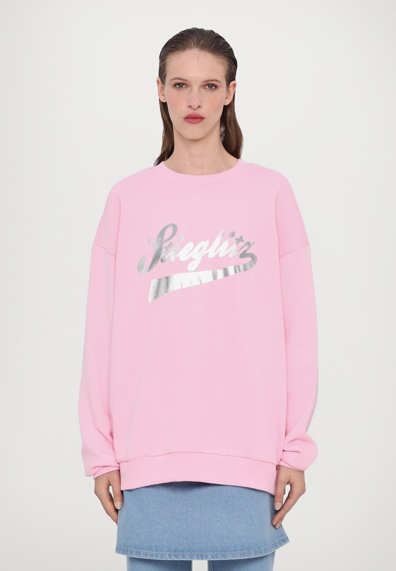Felpa oversized rosa con un design in lucido argento che recita "Steiglitz." Presenta un collo a giro e polsini a costine. Gonna di jeans mostrata sotto.