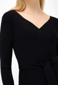 Svart omlottklänning i texturerad tyg med djup V-ringning, långa ärmar och knytet i midjan. Har ribbad detalj längs halsringningen.