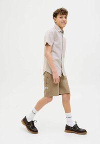 Chemise légère à manches courtes avec boutons, shorts beiges avec poches, chaussettes blanches, et chaussures brunes à semelles épaisses ; pose debout.