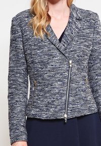 Námořnická modrá texturovaná bunda s těsným střihem, disponující zapínáním na zip vpředu, dvěma bočními kapsami na zip a nenápadnými kovovými akcenty.
