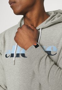 Sudadera gris con un logo impreso en azul claro y oscuro, con cordones blancos y una pequeña etiqueta de marca en el puño.