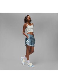 Ung kvinne står med hendene i lommene, iført lys grå shorts, blå treningsshorts, hvite sokker og blå-hvite sneakers.