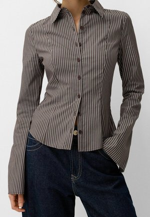 Mujer vistiendo una camisa ajustada de botones con rayas verticales marrones y blancas, de manga larga, y pantalones vaqueros azul oscuro.