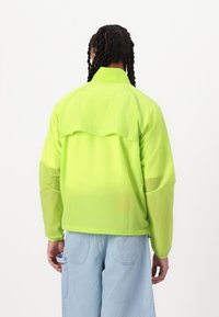 Neon green lightweight jacket with a relaxed fit, featuring a layered back design and long sleeves. Paired with light blue textured pants.