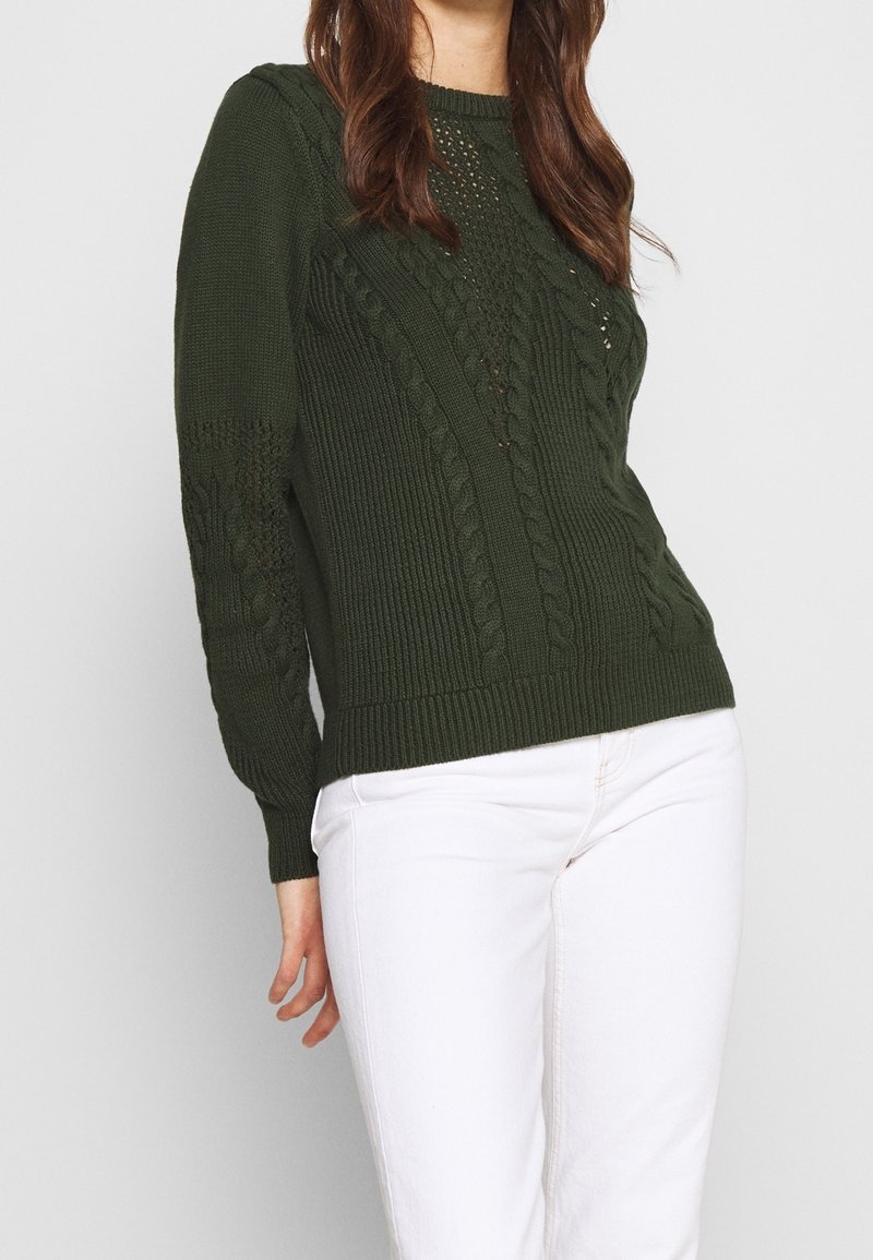 Pull en tricot vert foncé avec des poignets et un ourlet côtelés, présentant un motif de tricot en câble texturé et des détails de couture ouverte sur les manches.