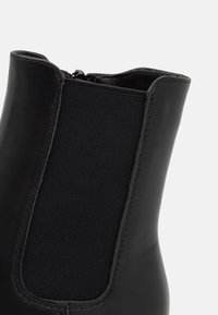 Bottine en cuir noir avec une finition lisse, un panneau latéral élastique et une fermeture éclair à l'arrière pour un enfilage facile.