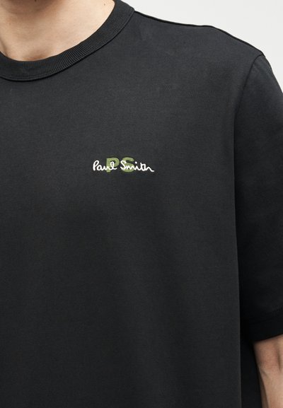 Μαύρο T-shirt με στρογγυλή λαιμόκοψη και μικρό πράσινο και άσπρο λογότυπο «Paul Smith» κεντημένο στο αριστερό στήθος, φορεμένο από ένα άτομο.