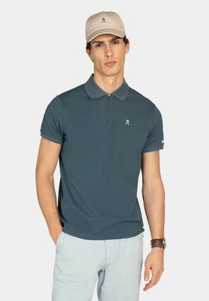 Hombre con camisa polo de manga corta color azul verdoso oscuro con cuello con cremallera, pantalones gris claro, gorra de béisbol beige y reloj de pulsera con brazalete.