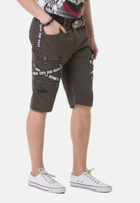 Khakifarbene Cargo-Shorts aus Baumwolle mit mehreren Reißverschlusstaschen. Mit weißem Text-Design und einem schwarzen Patch. Getragen mit weißen Sneakers.