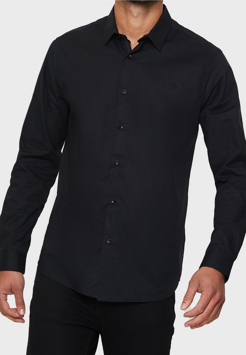Homme portant une chemise noire ajustée à manches longues avec col, debout devant un fond clair uni.
