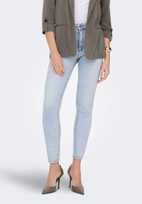 Άτομο που φορά στενά τζιν ανοιχτού γαλάζιου χρώματος, ταουπ γόβες με ψηλό τακούνι, λευκό τοπ και καφέ σακάκι με γυρισμένα μανίκια και χρυσό βραχιόλι.