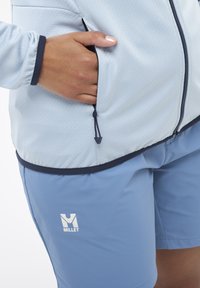 Hellblaues Jacket aus strukturiertem Stoff mit marineblauen Akzenten, ausgestattet mit Reißverschlusstaschen. Kombiniert mit hellblauen Shorts, Logo sichtbar.