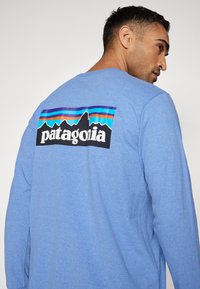 Hosszú ujjú kék póló, kontrasztos fekete grafikával, amely hegyeket ábrázol és "patagonia" felirat található a hátsó r�észén. Puha tapintású, laza stílusú.