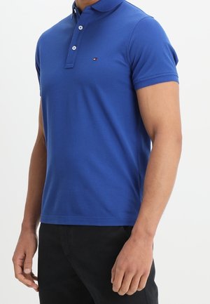 Mężczyzna ubrany w dopasowaną, krótką koszulkę polo w królewskim niebieskim kolorze, z dwoma białymi guzikami i małym logo na lewej piersi.