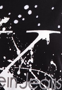 Tela negra con diseño de manchas de pintura blanca y una línea blanca inclinada parcial, con texto gris "calvin Jeans" en la parte inferior.