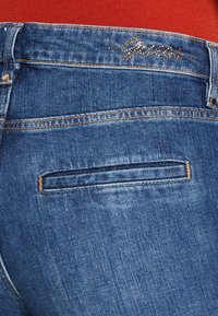 Dark blue denim jeans featuring a back pocket with a yellow stitch detail. The waistband displays the brand name "Guess" in metallic gold.