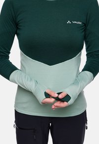 Person trägt ein zweifarbige grünes Langarmshirt mit Daumenlöchern, die Hände vor dem Körper zusammengelegt, und schwarze Hosen mit Reißverschlusstaschen.