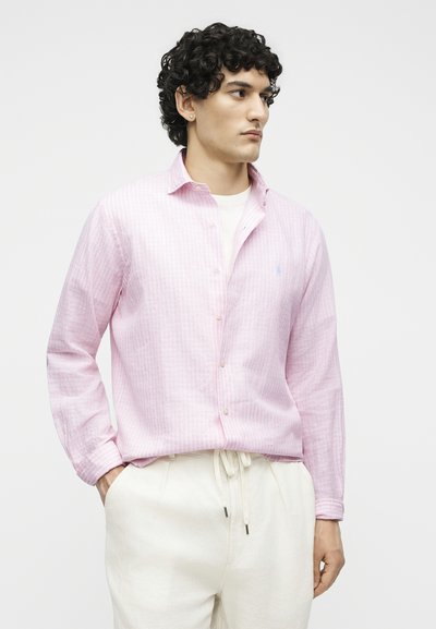 Άνδρας με κατσαρά μαλλιά που φοράει ανοιχτό ροζ ριγέ πουκάμισο με κουμπιά πάνω από λευκό τοπ και κρεμ παντελόνια, στέκεται με τα χέρια στις τσέπες.