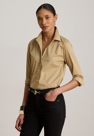 Femme aux cheveux foncés attachés en arrière, portant une chemise beige boutonnée avec des manches retroussées, un pantalon noir et une ceinture noire avec une boucle dorée.